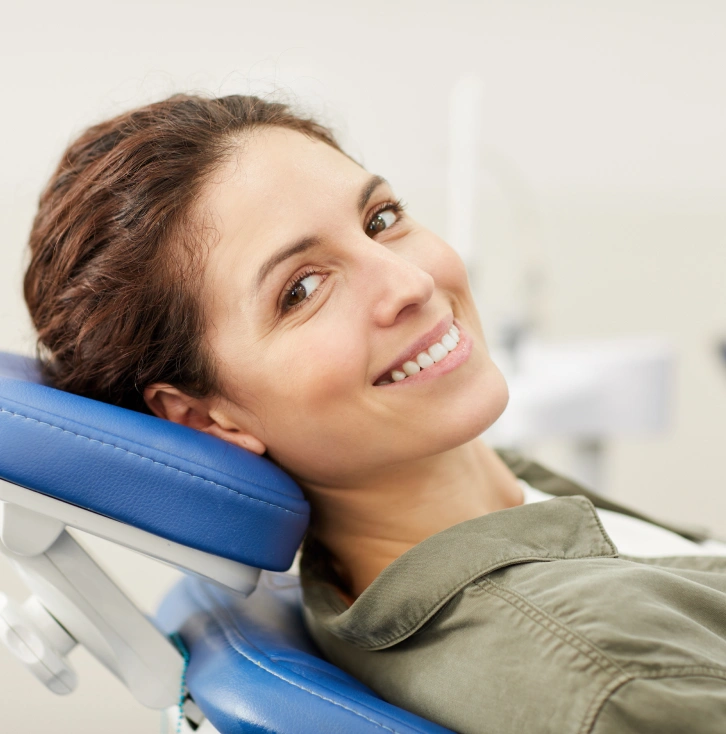 Woman smiling at dental office, holding blue neck pillow, clinical setting background.