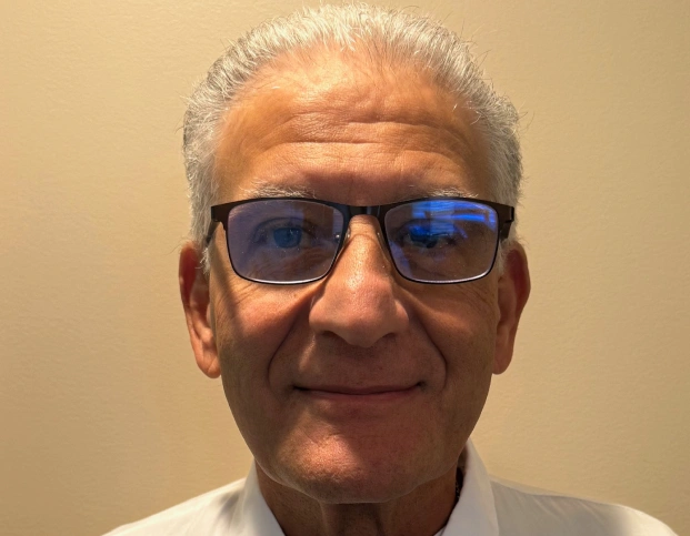 Older man wearing sunglasses and white shirt smiles at camera indoors.